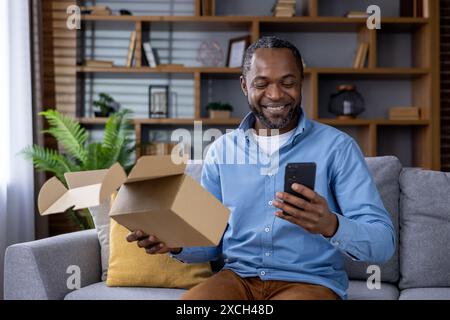Smiling man unboxing a package and using a smartphone while relaxing on the sofa at home, enjoying a delivery. Stock Photo