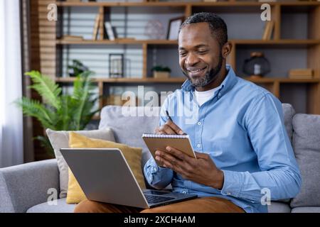 Smiling man taking notes while using a laptop in a cozy home office. Remote working, productivity, and home office concept. Stock Photo