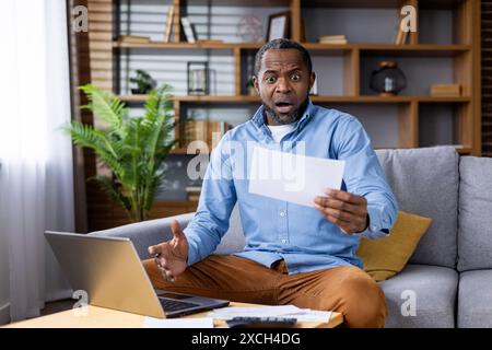 A surprised man holding a paper and looking at it with shock while sitting on a sofa and working on a laptop at home. Stock Photo
