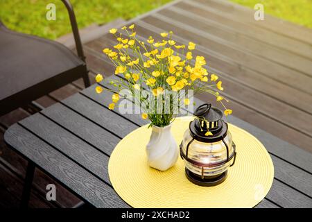 Bouquet of picked yellow Meadow buttercup or Ranunculus acris in a vase on home table, lantern with candle for decoration and yellow round place mat. Stock Photo