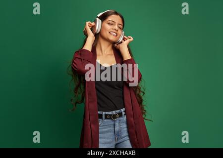 Overjoyed young Middle-eastern woman listening music in headphones and singing against green studio background. Stock Photo