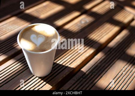 Paper cup with cappuccino with a heart shape , over square shaped shadows and sunshine pattern on a  wooden table Stock Photo