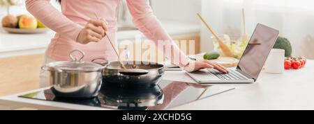 Woman Cooking Breakfast In A Modern Kitchen While Using Laptop Stock Photo
