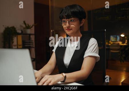 Young female coder in glasses working on laptop in dark office Stock Photo