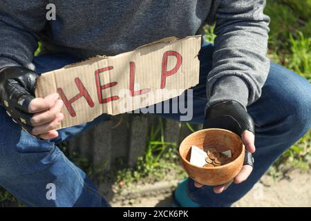 Poor homeless man holding help sign and bowl with donations outdoors, closeup Stock Photo