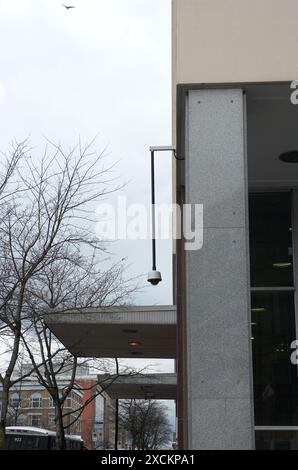 Surveillance camera on modern urban austere building exterior, with bird soaring above in overcast sky. Stock Photo