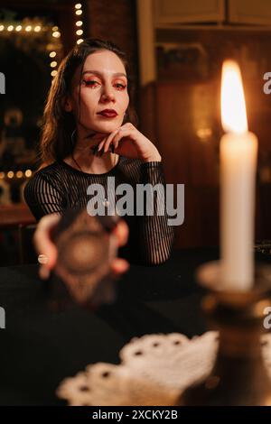 Vertical portrait of mysterious fortuneteller asking client to move deck of cards for layout during session, in dark esoteric room by light of burning Stock Photo