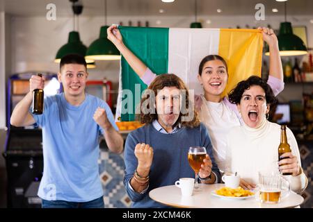 Company of emotional young adult fans supporting favorite team with flag of Ireland while resting in pub with beer Stock Photo