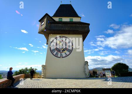 A clock tower with a large clock face and a small clock on the side. The tower is tall and has a steeple. There are several people walking around the Stock Photo