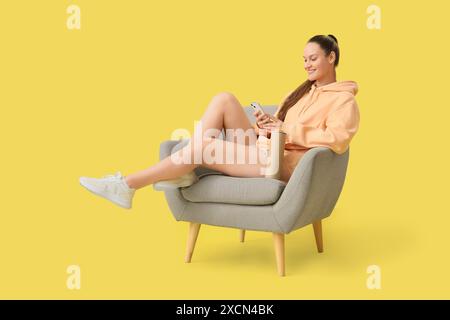 Young woman with potato chips using mobile phone in armchair on yellow background Stock Photo