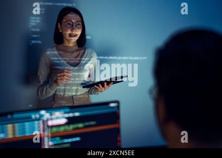 asian woman developers collaborating on a coding project, with one member presentation at code projected on the wall while the other works on multiple Stock Photo