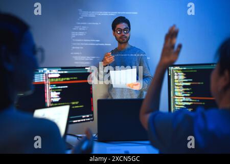 developer conducts a code review meeting, pointing at projected code on the wall , with colleagues engaging and asking questions Stock Photo