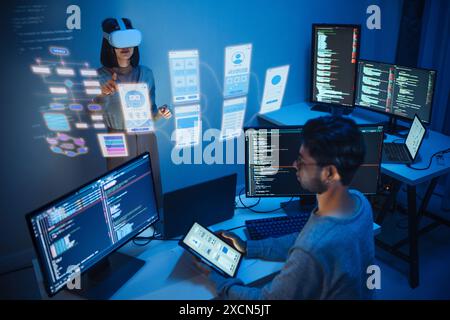 developer uses a VR headset to interact with projected interface designs, while colleagues discuss and code on screens at office Stock Photo