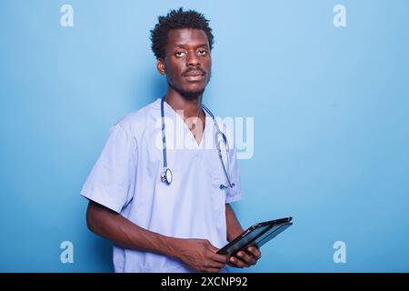 African American doctor in scrubs holds medical devices, including a digital tablet. Against blue background, male nurse is ready to provide healthcare with modern technology. Stock Photo