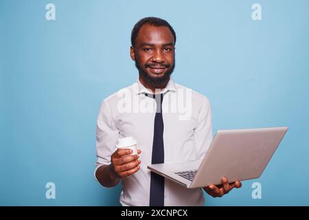 African american writer looking at camera while carrying digital laptop and cup of coffee on blue background. Male entrepreneur casually using wireless computer while having a beverage. Stock Photo