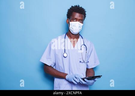 African American doctor in blue scrubs and face mask stands against a blue background while holding a tablet. Black man with stethoscope and gloves, grasping a digital device. Stock Photo