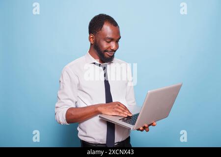African american male blogger confidently holding laptop, looking at the wireless portable device with a smile. Young entrepreneur in video call conference on minicomputer. Stock Photo