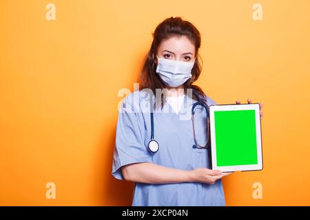 Healthcare specialist vertically holding tablet with green screen display on her left side. Female nurse wearing face mask is gripping a device with blank chromakey copyspace template. Stock Photo