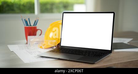 table architectural blueprints and hardhat laptop on a desk in an office setting, displaying a blank screen, representing technology and work Stock Photo