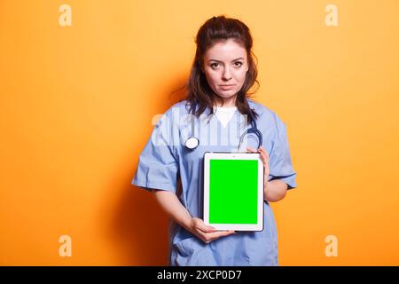 Healthcare professional vertically holding tablet with green screen. Woman wearing nurse uniform and stethoscope is grasping digital device displaying blank chromakey mockup template. Stock Photo