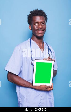 African American male nurse in scrubs standing against blue background with digital device showing green screen. Black man vertically grasping a tablet with chromakey mockup template. Stock Photo