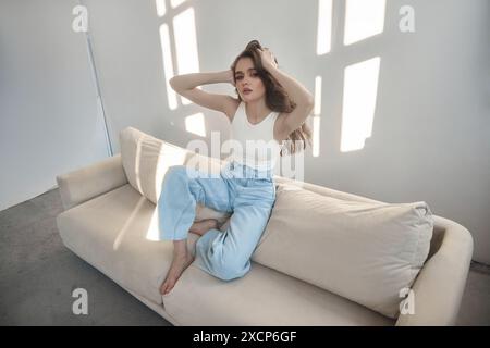 Young beautiful woman with long dark hair sits on white sofa, running her fingers through hair. Room is bright and sunny, with light streaming in thro Stock Photo