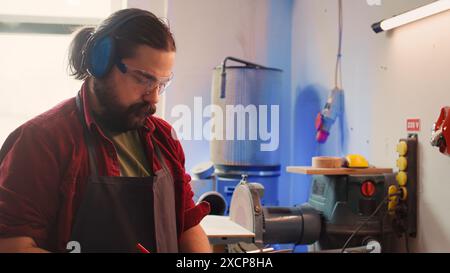 Craftsperson at work consulting blueprint and using orbital sander with sandpaper on lumber. Woodworking expert in carpentry shop uses angle grinder on wood after checking schematics, camera B Stock Photo