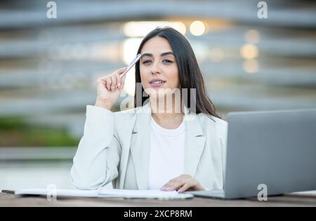 Woman Uses Pen to Think While Working on Laptop Outdoors Stock Photo