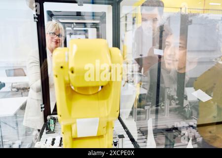 Engineers closely examining a yellow robotic arm's functionality during a hands-on training session in an industrial environment. Stock Photo