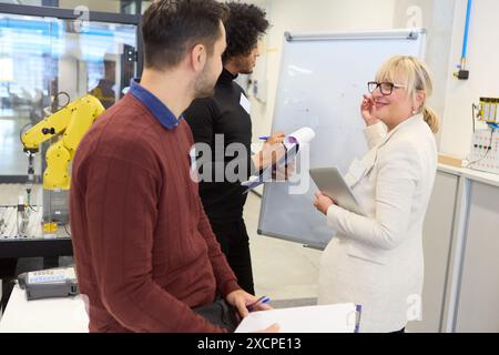 Team of engineers collaborating on programming and automation for robotics in an industrial environment with a robotic arm in the background. Stock Photo