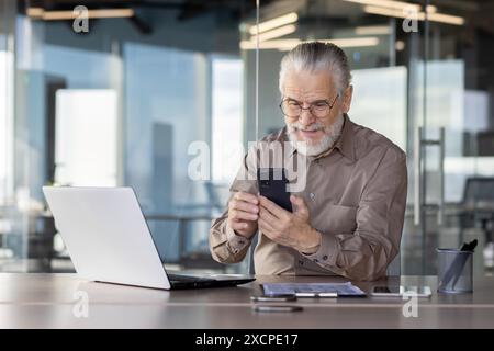 Senior businessman sitting at office desk using smartphone and laptop, working and communicating. Stock Photo