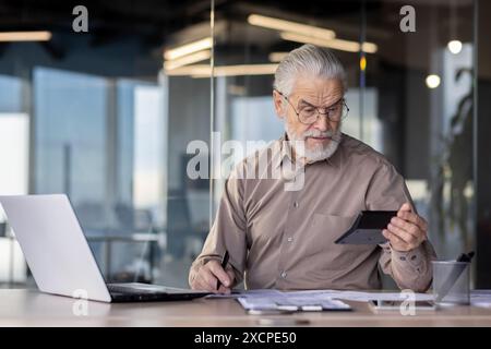 Elderly man in a modern office environment, using a laptop and tablet for work. Professional business setting. Stock Photo