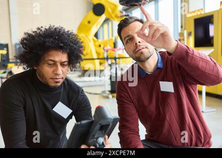 Two engineers collaborate on programming a robotic arm in an industrial environment, focusing on automation and advanced technology. Stock Photo
