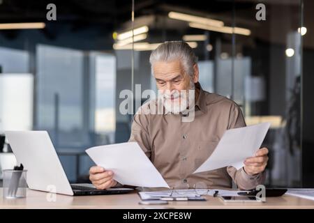 Senior businessman in a modern office reviewing paperwork at his desk with a laptop and reading glasses Stock Photo