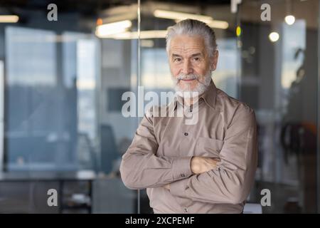 Confident senior businessman standing with arms crossed in a modern office setting, looking at camera. Stock Photo