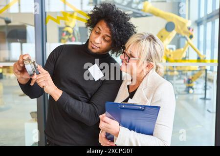 Two engineers discussing a robotics component in an industrial setting, with robotic gripper arms visible in the background. Focus on collaboration an Stock Photo