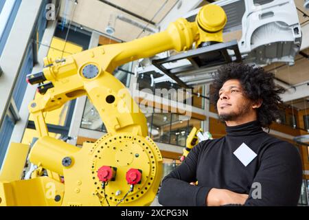 Engineer standing confidently in front of a yellow industrial robot arm in a contemporary factory environment, indicating technological advancement an Stock Photo