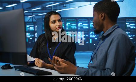 Coworkers in data center looking over annual systems review documents, discussing about parameters. Colleagues doing brainstorming in high tech workspace, talking about server clusters specs Stock Photo
