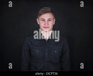 Portrait of a carefree young man against a black wall background looking to camera. Relaxed male model with joyful face expression wearing dark blue j Stock Photo