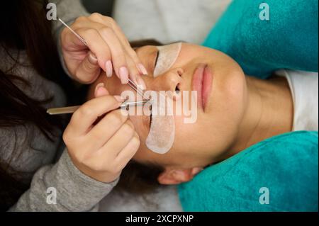 Close-up of a woman receiving eyelash extensions in a beauty salon. The beautician is using tweezers to apply individual lashes. Stock Photo