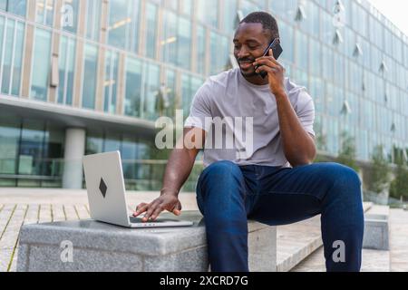 African American man freelancer using laptop talking on phone on urban street in city. Business man having virtual meeting. Professional programmer using phone laptop working outdoors. Remote work Stock Photo