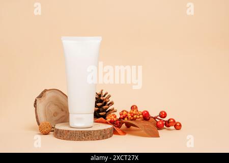 a white cosmetic tube placed on a wooden slice surrounded by pine cones, berries, and autumn leaves against a beige background Stock Photo