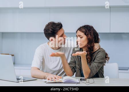 Couple sits at table, deeply engaged in serious financial discussion. Man tries to comfort woman, who looks frustrated and helpless. Laptop, notebook, and calculator are on table, emphasizing stress and concern over their finances. Stock Photo