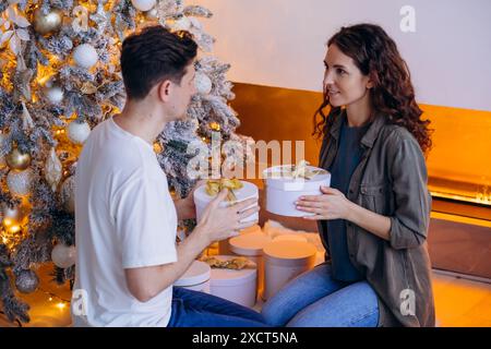 Young couple is seen exchanging gifts while sitting by beautifully decorated Christmas tree with golden and white ornaments. Scene is cozy and festive, with warm lighting and fireplace in background.  Stock Photo