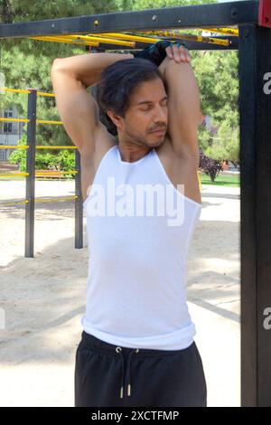 A man with a blue wristband is standing in front of a metal structure. He is wearing a white tank top and black shorts Stock Photo