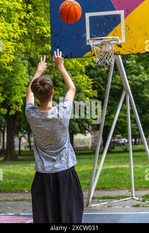 A young man is seen from behind as he shoots a basketball towards the hoop on an outdoor court. The basketball is in mid-air, and the player is reachi Stock Photo