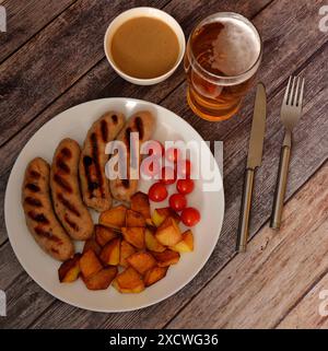 Grilled sausages with fried potatoes and cherry tomatoes on a plate, next to mustard sauce and a glass of light beer on a wooden table. Top view, flat Stock Photo