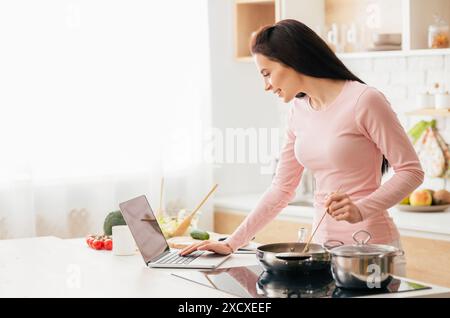 Woman Standing in Kitchen Using Laptop, Reading Recipe Stock Photo