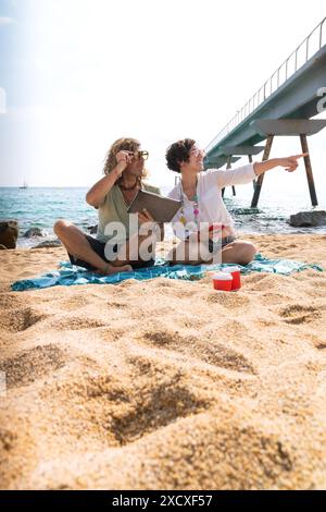 Couple on the beach,pointing to the location of the hotel with the tablet.Vertical Stock Photo