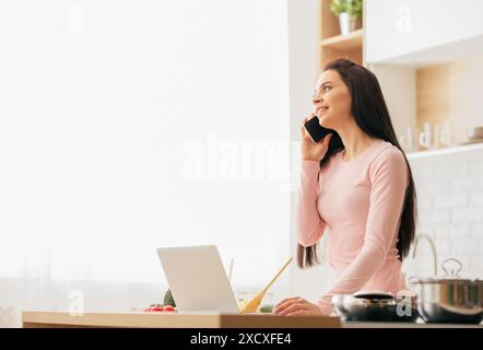 Woman Talking on Phone While Using Laptop, Kitchen Interior Stock Photo
